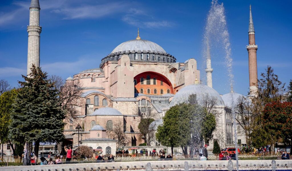 Exterior view of Hagia Sophia in Istanbul, Turkey, showcasing its massive central dome, surrounding minarets, and a foreground fountain with visitors enjoying the sunny day.