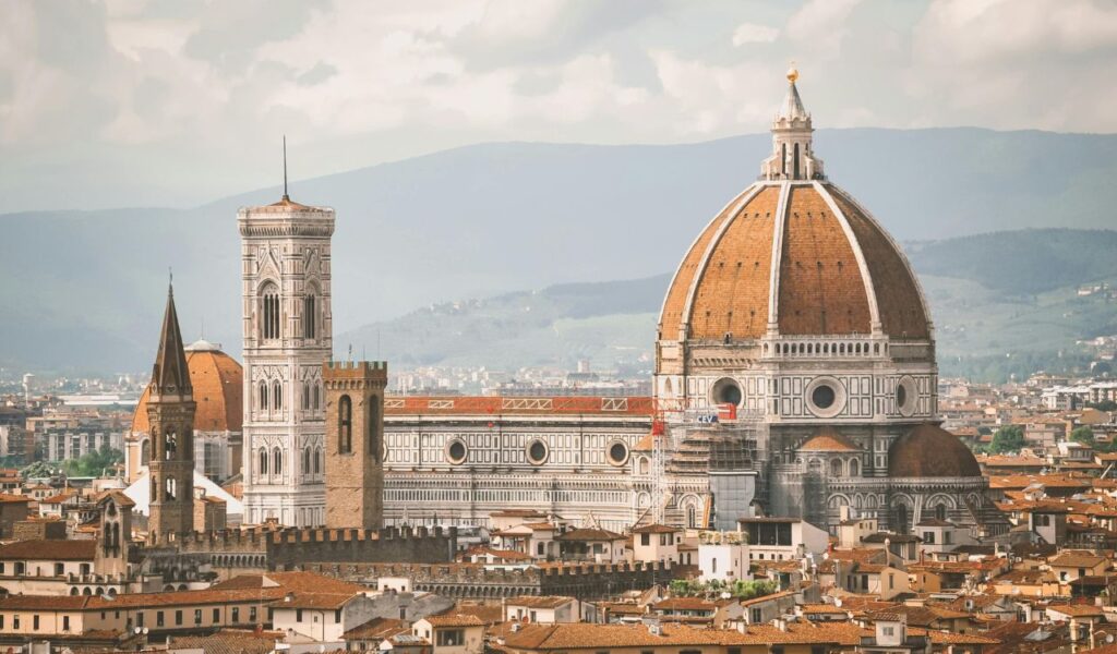 Panoramic view of Florence Cathedral in Italy, featuring its iconic red-tiled dome, Giotto’s bell tower, and surrounding historic city rooftops with rolling hills in the background.