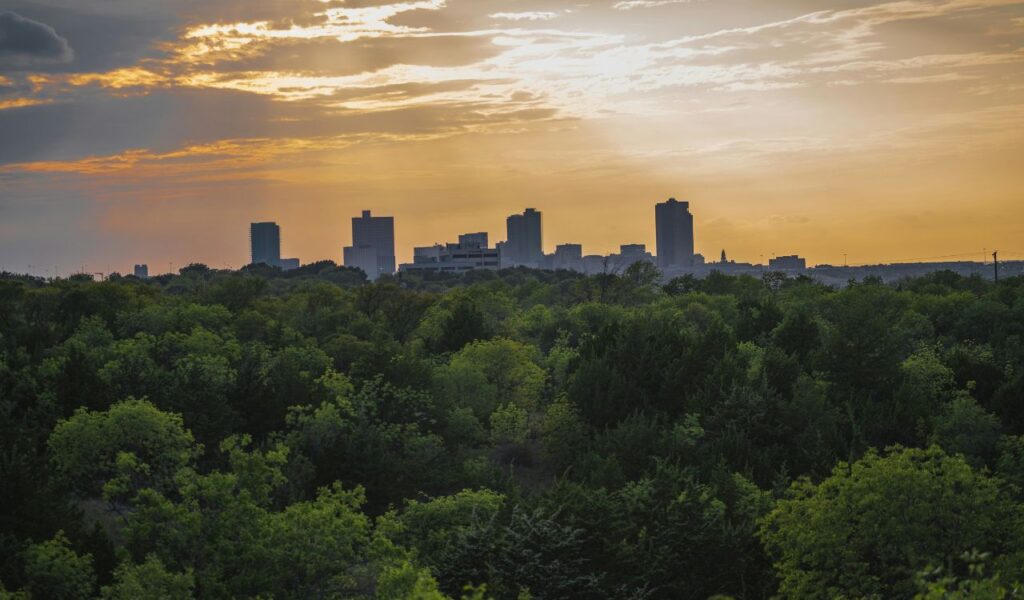 Silhouette of Fort Worth, Texas skyline at sunset with dense green trees in the foreground and a glowing orange sky.