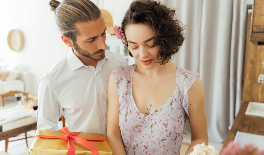 A man in a white shirt presents a gift to a smiling woman in a floral dress, sharing a tender moment that reflects appreciation and genuine compliments.