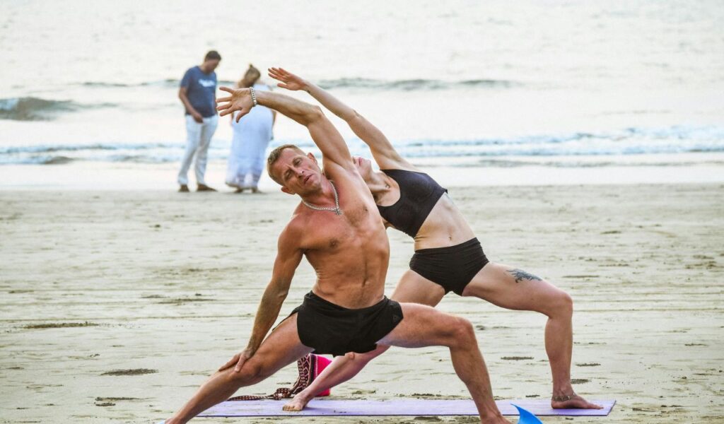 Two people practicing a yoga pose together on a sandy beach in Goa, India, with the ocean waves and other beachgoers in the background.