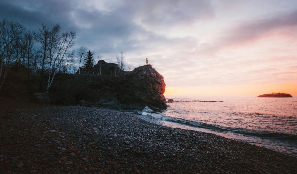 A person in a yellow jacket stands atop a rocky cliff at Grand Portage National Monument in Minnesota, overlooking Lake Superior during a colorful sunrise, with gentle waves lapping at the pebble-covered shore.