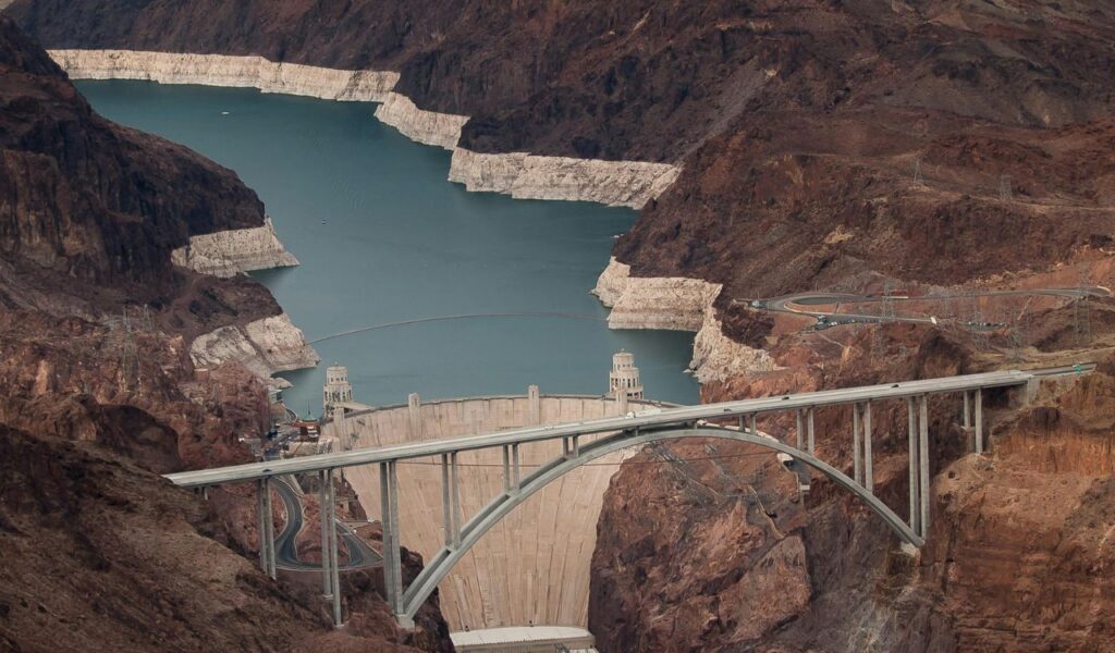 A wide view of the Hoover Dam surrounded by rocky canyons and the Colorado River, showing the bridge and water reservoir in the distance.