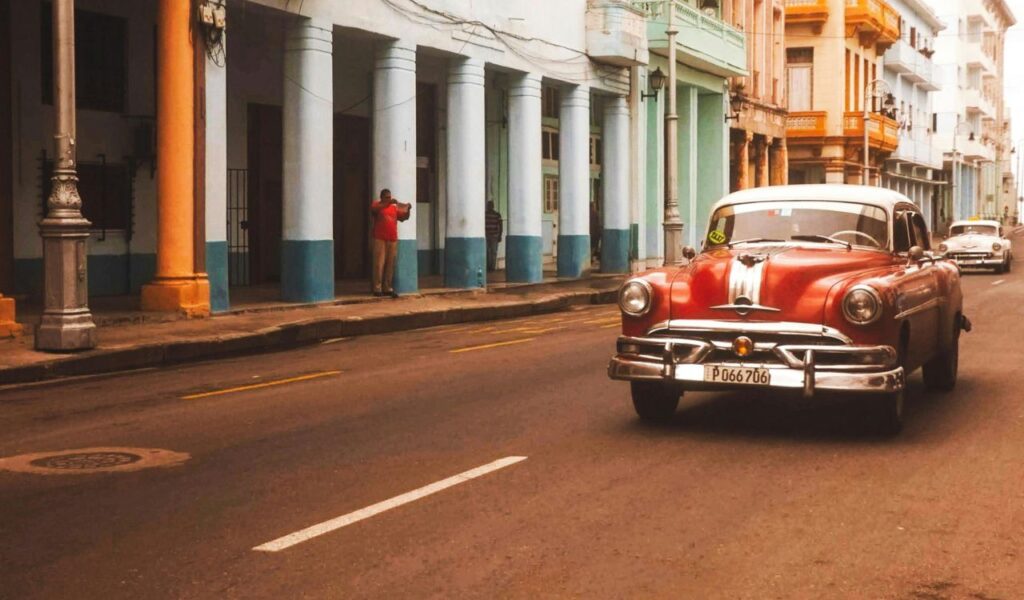 Classic red vintage car driving through the colorful colonial streets of Havana, Cuba, with pastel buildings and balconies in the background.
