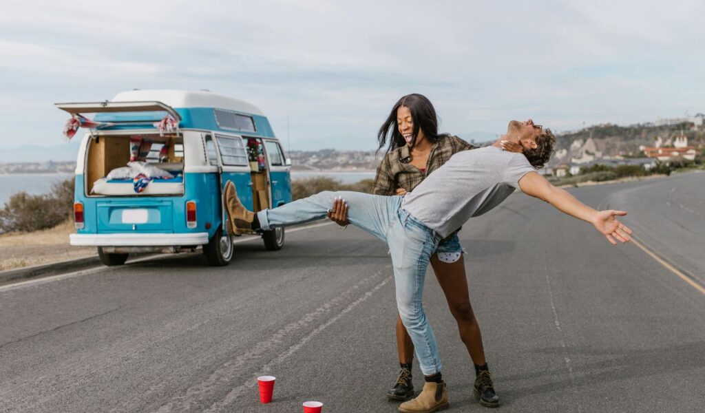 A cheerful couple playing together on an empty road with a blue camper van parked nearby, laughing and enjoying a carefree moment during a road trip.