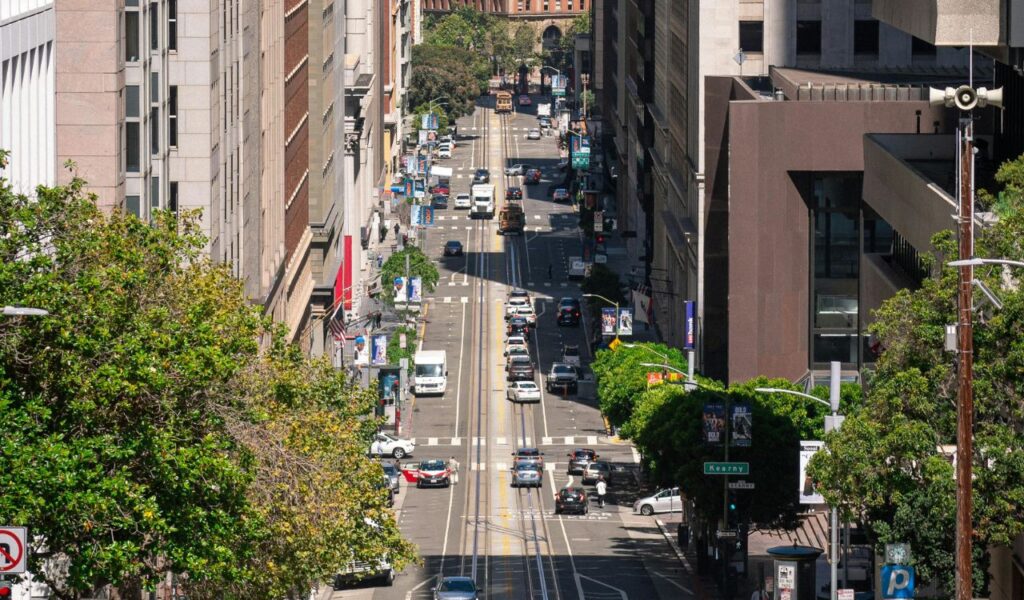 usy urban street in San Francisco with cars, pedestrians crossing, and the Bay Bridge visible in the background on a sunny day.