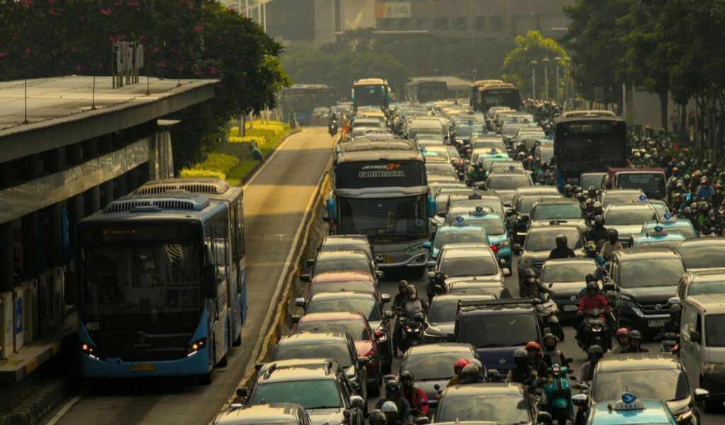 Heavy traffic congestion with cars, buses, and motorcycles crowding multiple lanes on a busy urban road lined with trees and buildings.