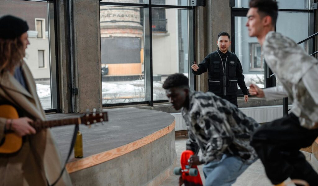 A group of young people are indoors, one holding a guitar and another with a skateboard, while a man approaches them, appearing to intervene.