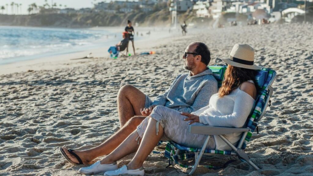 A couple sitting on beach chairs facing the ocean, relaxing on a sandy beach with a few people and coastal houses in the background.