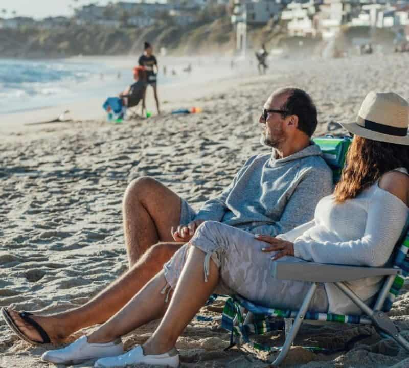 A couple sitting on beach chairs facing the ocean, relaxing on a sandy beach with a few people and coastal houses in the background.