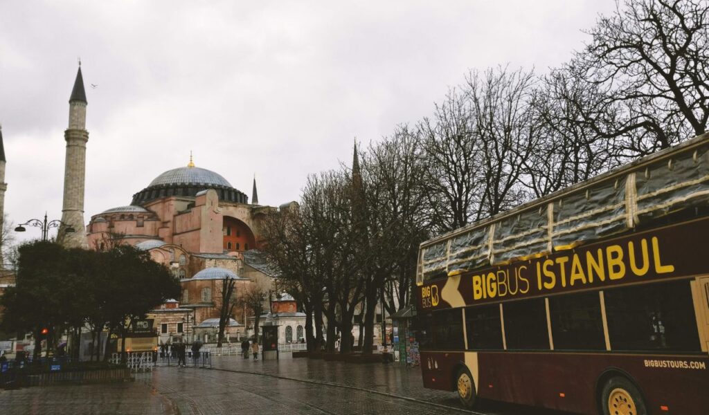 A sightseeing bus labeled "Big Bus Istanbul" passes by the historic Hagia Sophia with its domes and minarets on a cloudy day in Istanbul, Türkiye.