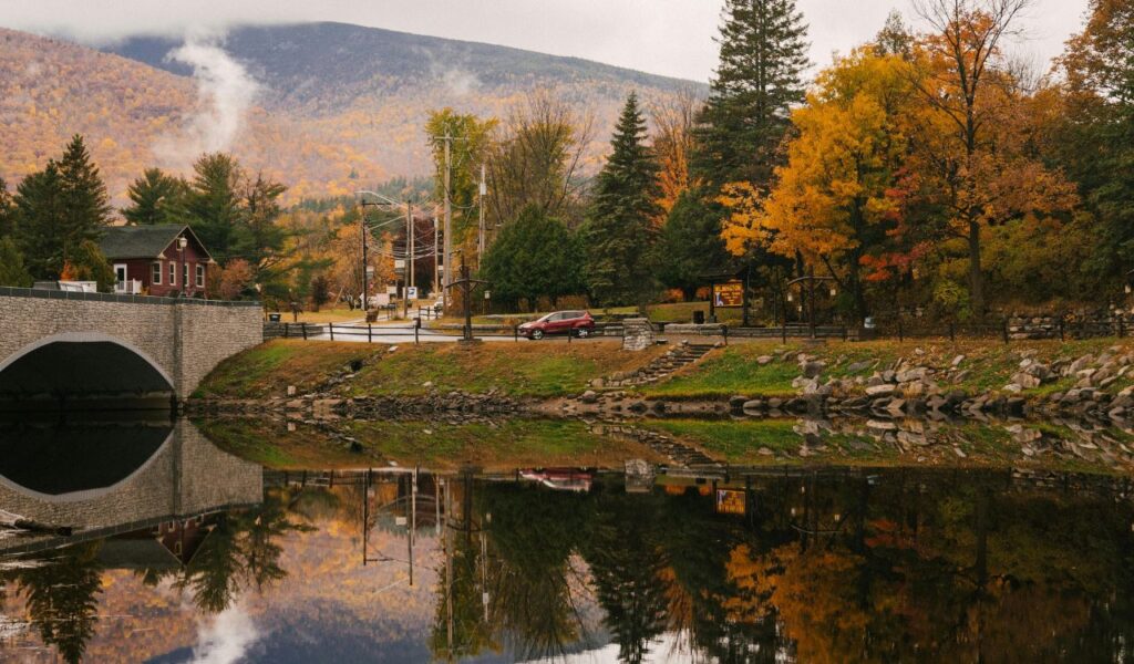 A serene autumn view of Jaffrey, New Hampshire, with colorful foliage, a stone bridge, and still waters reflecting the trees and nearby mountains.