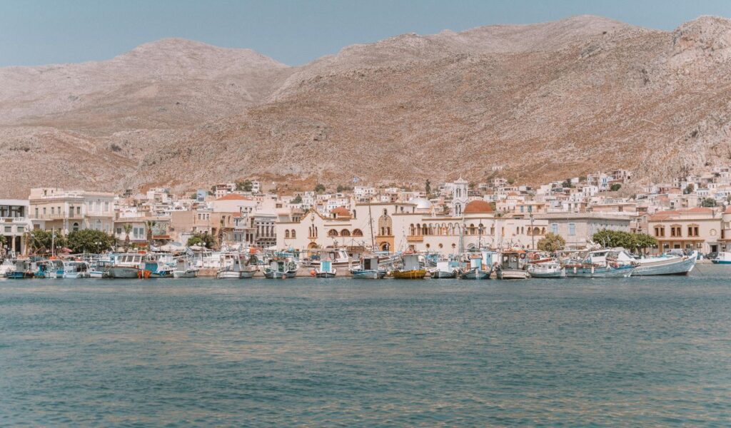 Fishing boats line the harbor of Kalymnos, Greece, with neoclassical buildings and hillside houses set against a backdrop of rugged mountains.