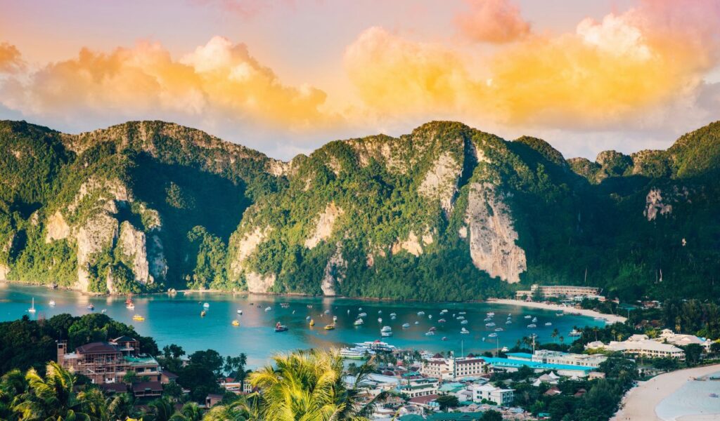 t: A vibrant coastal town nestled between two lush green cliffs in Thailand, with turquoise waters dotted by colorful boats and a golden sunset sky glowing above.