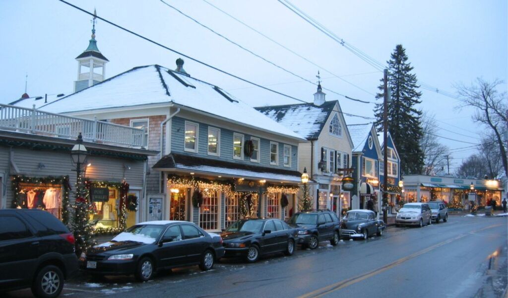 A cozy winter scene in Kennebunkport, Maine, with snow-dusted rooftops, holiday lights, decorated storefronts, and parked cars lining a quaint street.