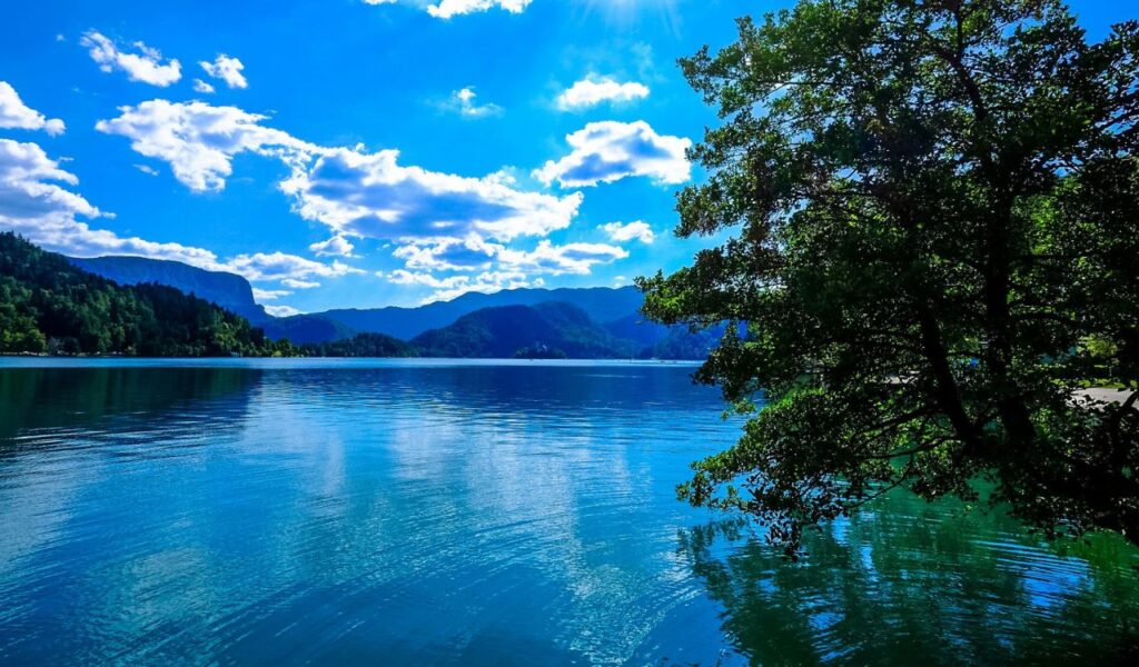 Crystal-clear waters of Lake Bled, Slovenia, with surrounding green hills, distant mountains, and a bright blue sky dotted with clouds.