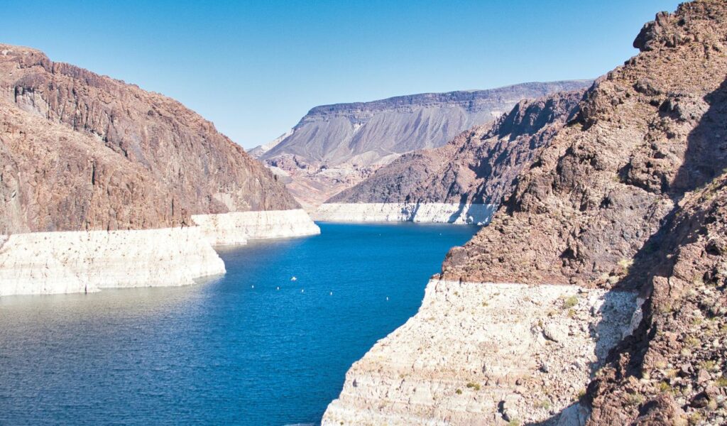 View of Lake Mead surrounded by rocky cliffs with visible waterlines, showing the effects of receding water levels under a clear blue sky.