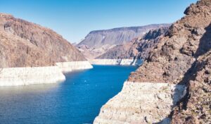 View of Lake Mead surrounded by rocky cliffs with visible waterlines, showing the effects of receding water levels under a clear blue sky.