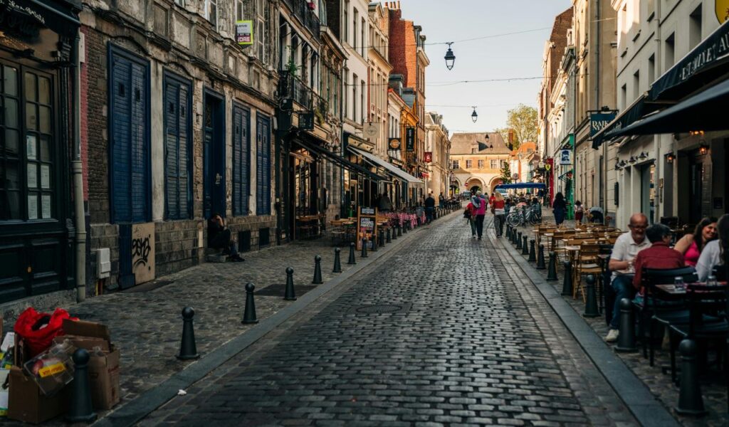 Cobblestone street in Lille lined with historic buildings, outdoor cafés, and people walking under a clear evening sky.