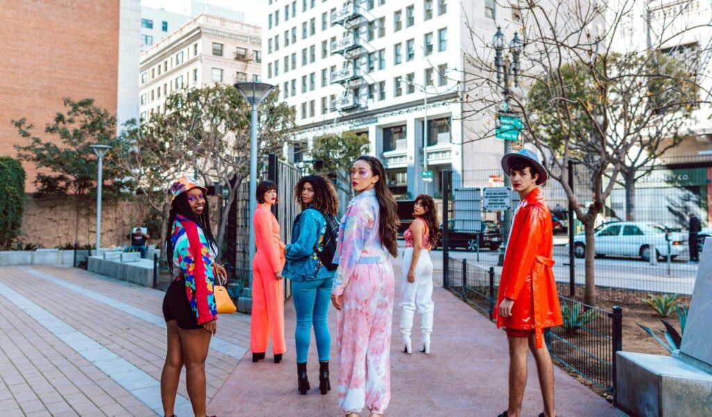 A group of stylish young adults in colorful outfits stands on a city sidewalk with mid-rise buildings in the background.