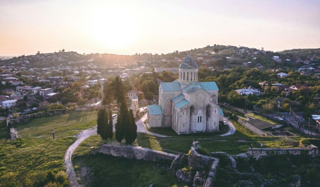 A historic stone church with teal roofs stands on a hill in Macon, Georgia, surrounded by green trees and homes under a soft evening sun.