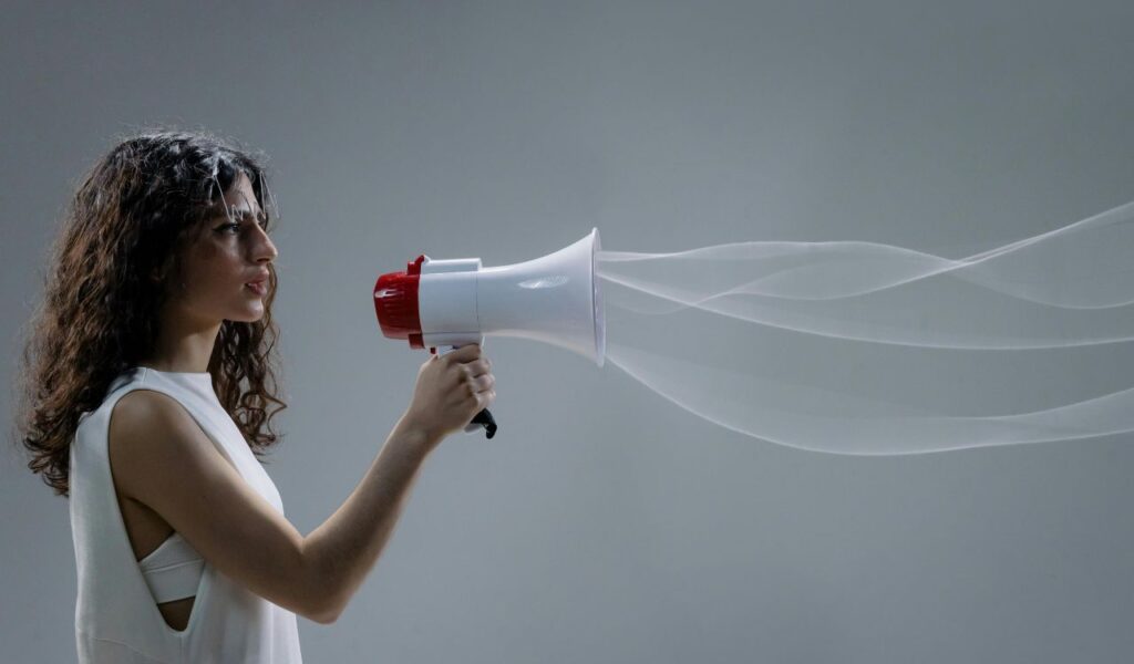 A woman with curly hair holds a red and white megaphone, speaking into it as illustrated sound waves extend outward against a gray background.