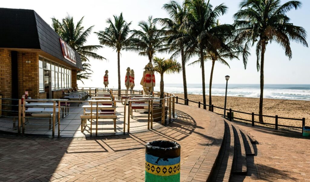 Outdoor café terrace with empty tables facing a sandy beach, lined with palm trees, on a sunny day by the ocean.
