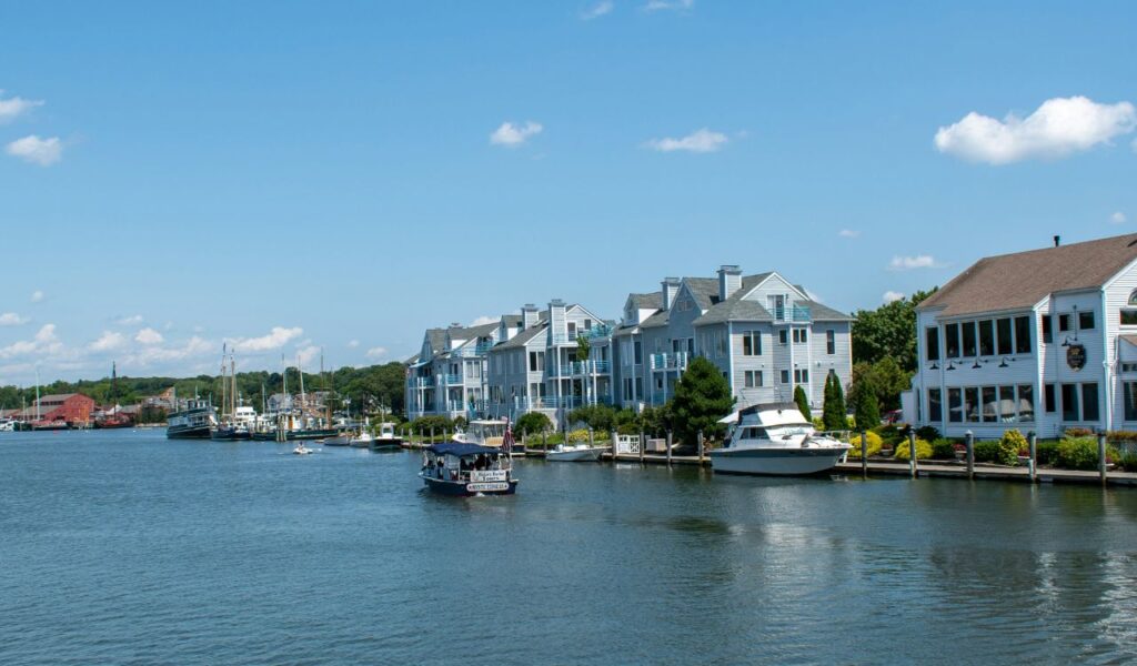 A sunny waterfront scene in Mystic, Connecticut, with boats cruising along the calm river and pastel-colored buildings lining the scenic shoreline.