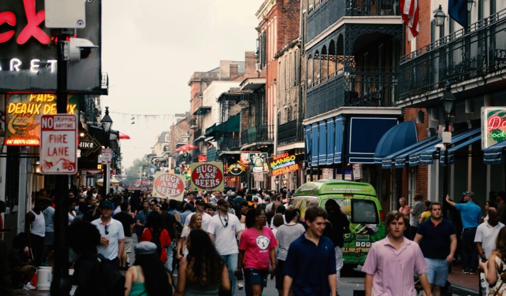 Crowds walk along Bourbon Street in New Orleans, lined with colorful signs, balconies, and neon-lit bars during a festive evening.