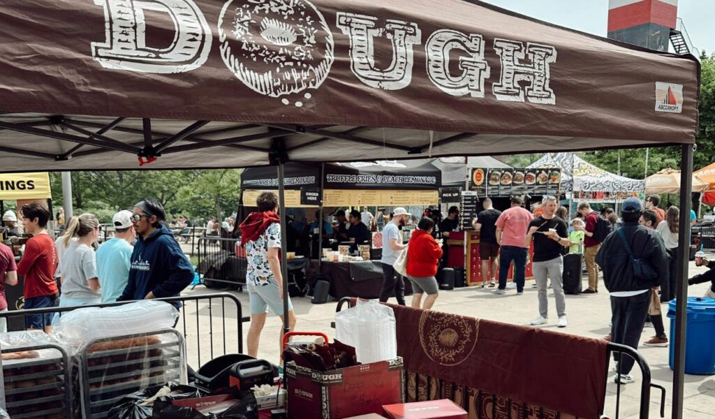People gather at an outdoor food market in New York City, with vendor tents offering various dishes, including a stand labeled “DOUGH.”