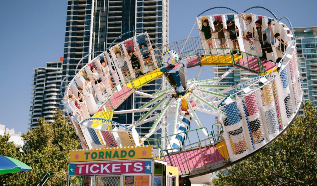 A colorful carnival ride called “Tornado” spins high in the air with riders, set against a backdrop of tall modern buildings and clear skies.