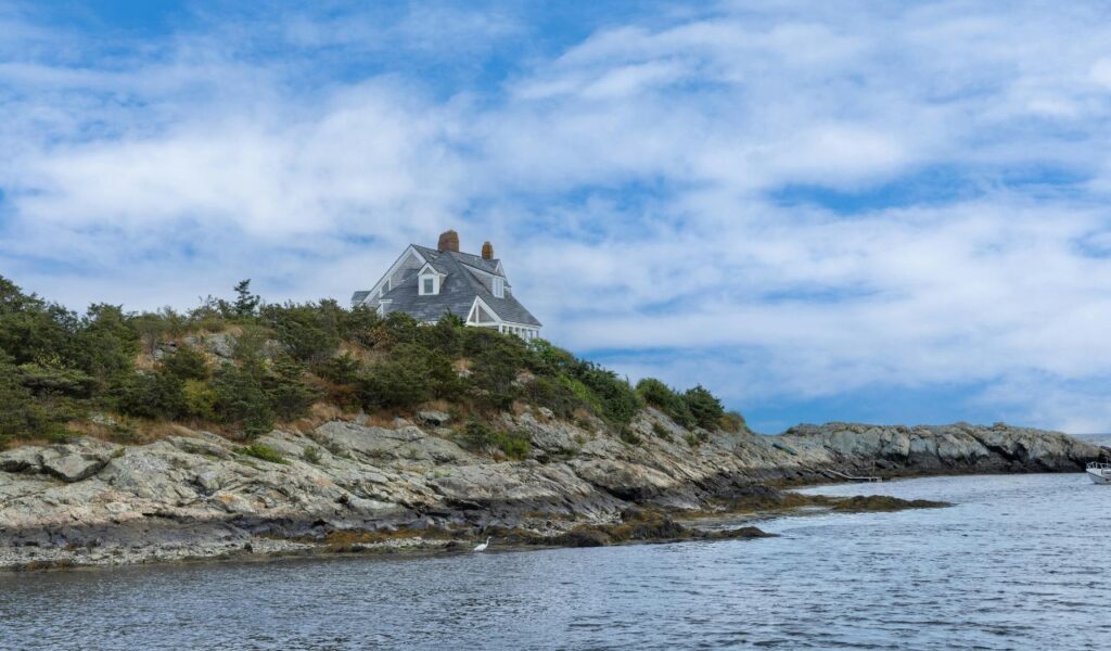 A charming coastal house perched atop a rocky shoreline in Newport, Rhode Island, surrounded by lush greenery under a partly cloudy blue sky.