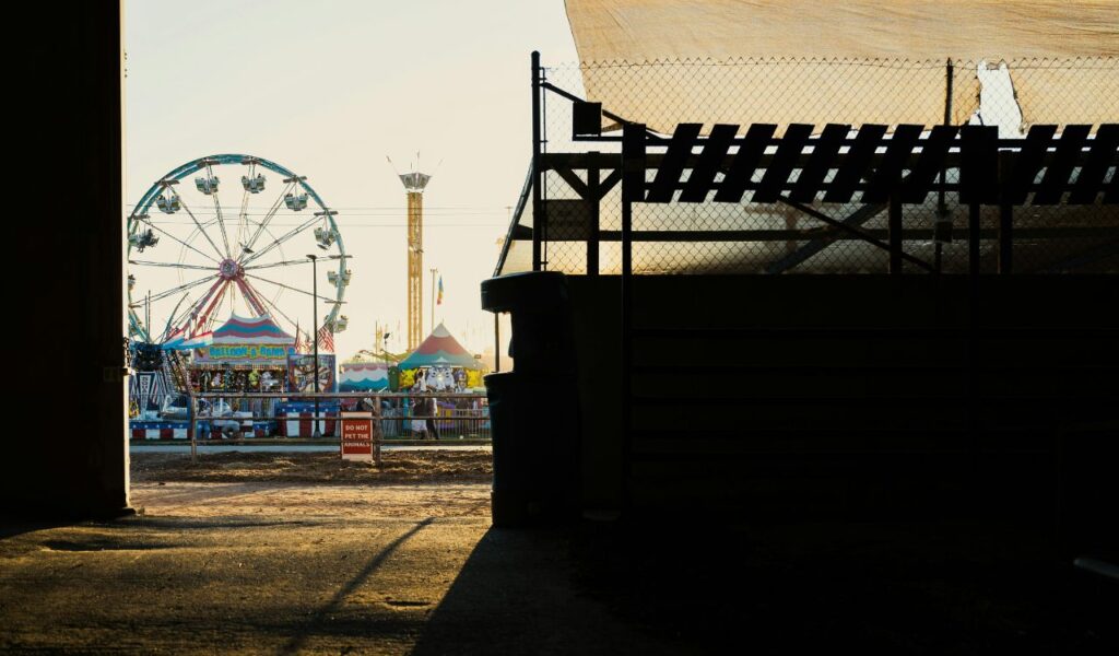 View of a fairground through a shaded entryway, featuring a Ferris wheel, colorful tents, and rides at sunset, with a “Do Not Pet the Animals” sign in the foreground.