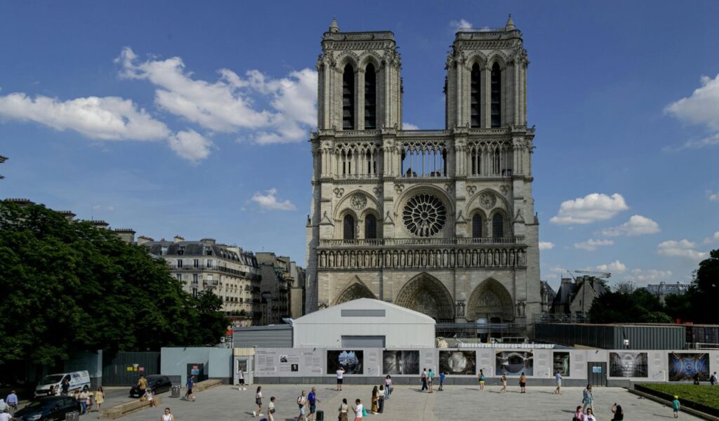 The front facade of Notre Dame Cathedral in Paris, France, with its twin towers, rose window, and Gothic architecture, surrounded by restoration work and visitors walking in the square below.