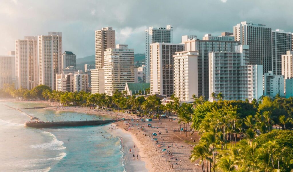 Aerial view of Waikiki Beach in Oahu, Hawaii, with turquoise waters, sandy shoreline, palm trees, and tall hotels lining the coast.