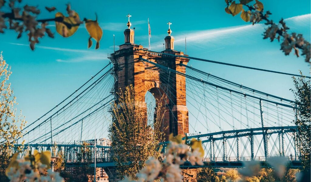 altext ChatGPT said: Framed by blooming branches, the John A. Roebling Suspension Bridge spans the Ohio River, connecting Cincinnati, Ohio, to Covington, Kentucky, under a clear blue sky.