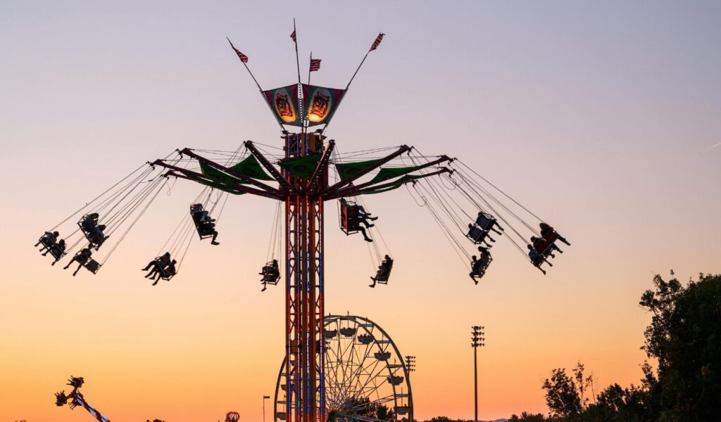 People enjoy a high-flying swing ride at a state fair during sunset, with a Ferris wheel and other attractions in the background.