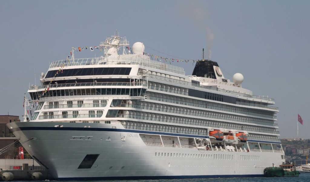 A large white cruise ship docked at a port with multiple decks, lifeboats, and colorful flags strung across the top under a clear sky.