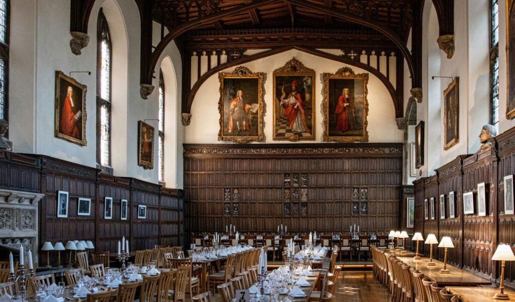 Elegant historic dining hall in Oxford with long wooden tables, ornate paneling, high arched ceiling, and portraits on the walls.