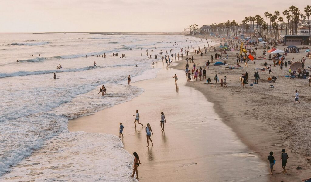 A crowded beach with people walking along the shoreline, swimming in the ocean, and relaxing on the sand under umbrellas during sunset.