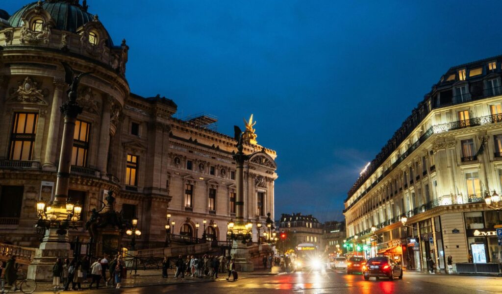 Evening street scene in Paris with the illuminated Palais Garnier opera house, bustling traffic, and pedestrians under a deep blue sky.








