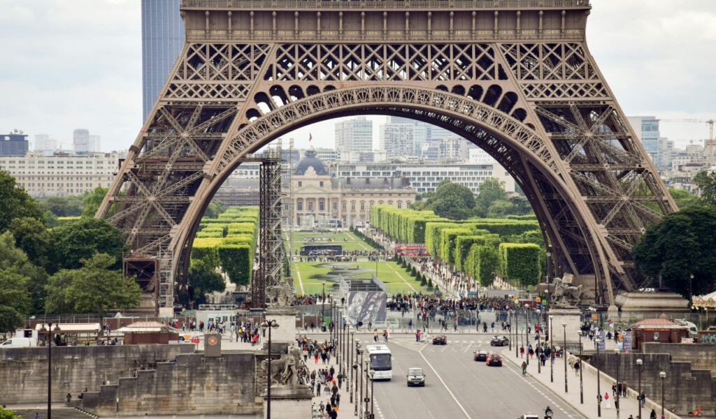 View from beneath the Eiffel Tower in Paris, showing its iron framework, tree-lined Champ de Mars gardens, and bustling streets filled with people and vehicles.