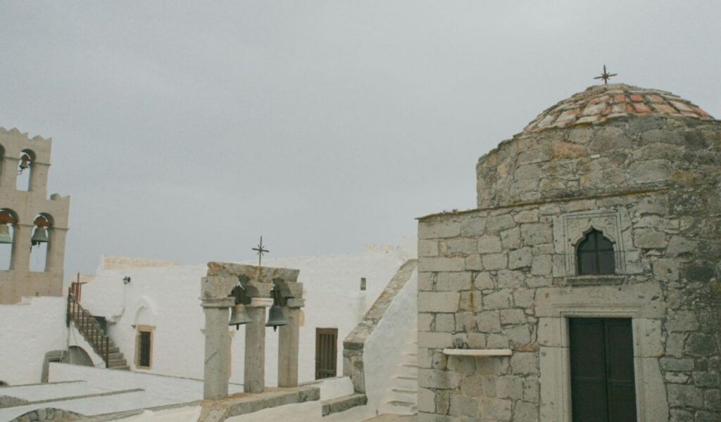 A small whitewashed chapel with a blue door and bell stands by the rocky coastline of Amorgos, Greece, overlooking the deep blue Aegean Sea.