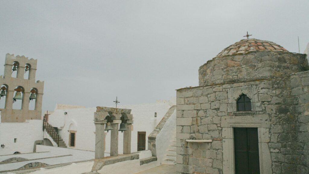 Historic stone buildings and bell towers of the Monastery of Saint John on Patmos, Greece, stand against a cloudy sky.