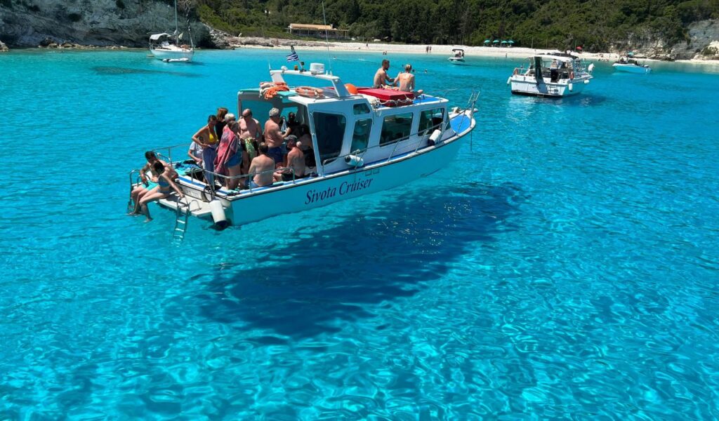 A small tour boat filled with people floats on the crystal-clear turquoise waters off Paxos, Greece, with a sandy beach and green hills in the background.