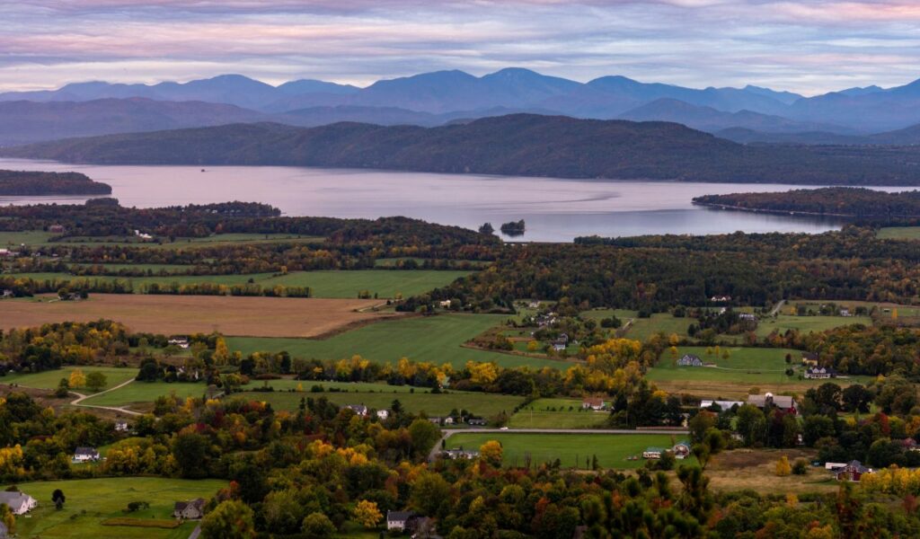 A panoramic view of Peacham, Vermont, showing rolling farmlands and scattered homes, with Lake Champlain and the Adirondack Mountains in the distance.