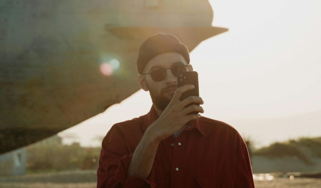 : A man wearing sunglasses and a beanie takes a photo or video with his smartphone outdoors near a large structure during sunse