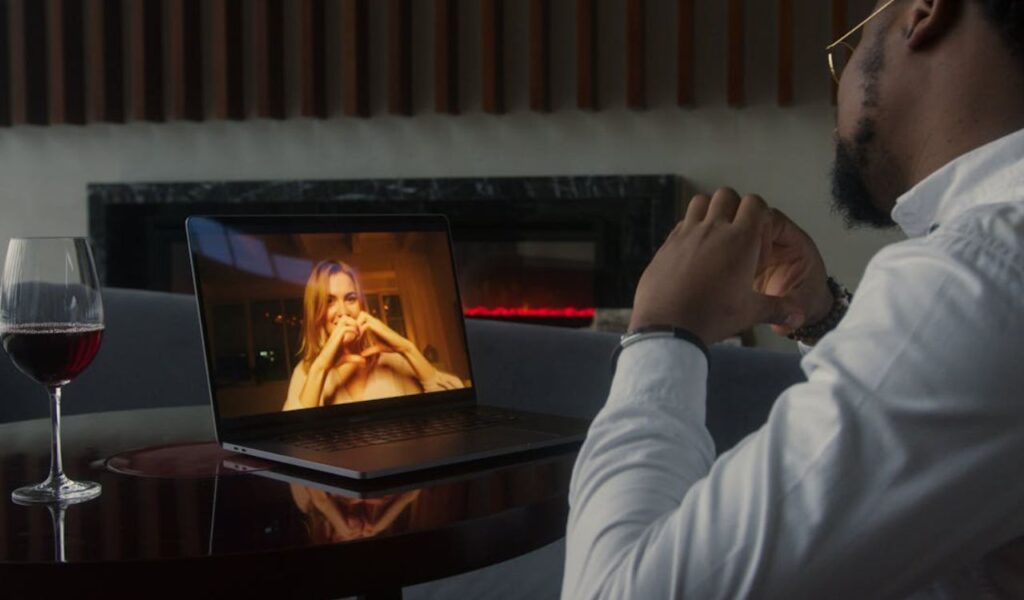 A man sits at a table with a glass of red wine, making a heart shape with his hands toward a woman on a laptop screen who is smiling and returning the gesture during a video call.