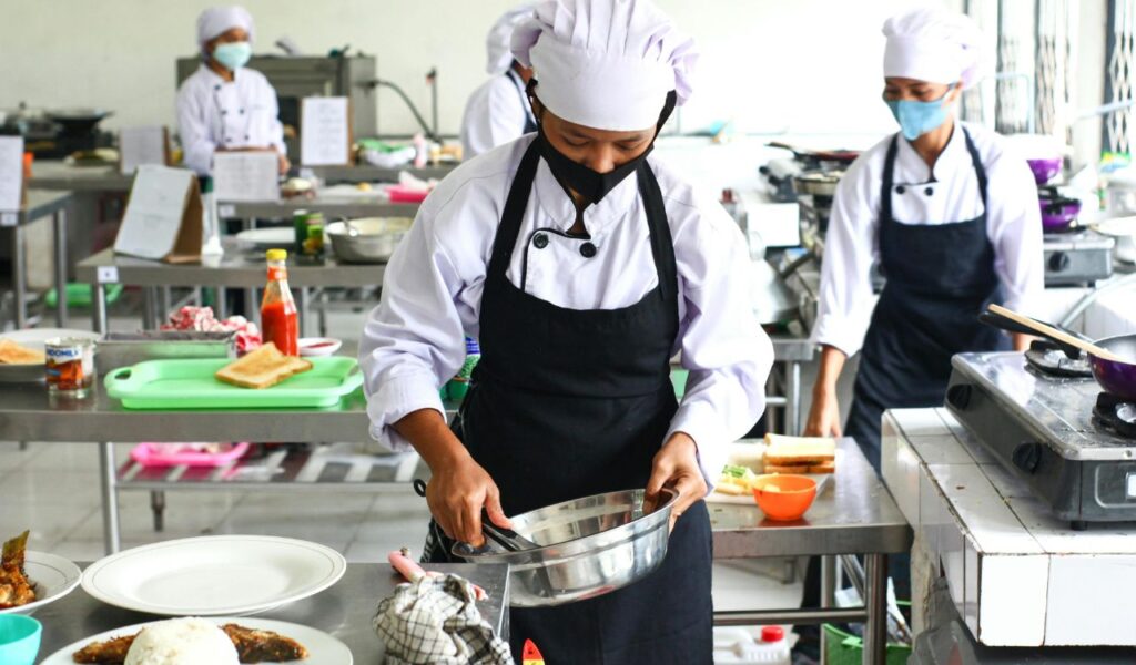 Chefs wearing clean uniforms and masks prepare food in a commercial kitchen, following proper food handling and hygiene practices.