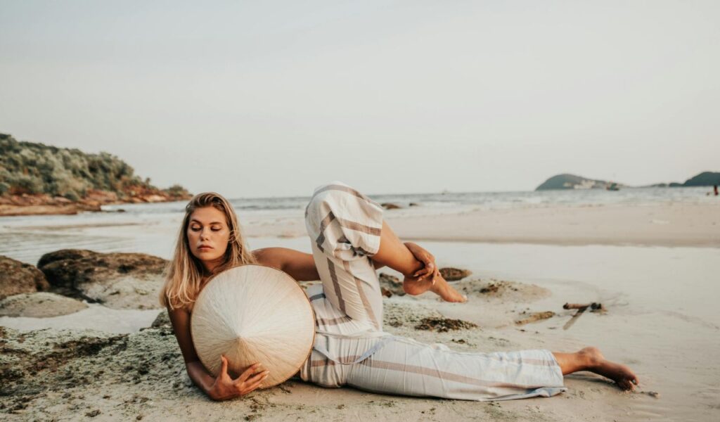 A woman practicing yoga on a serene beach at low tide, holding a conical Asian hat while lying on the sand near rocks, with calm ocean waves and distant islands in the background.